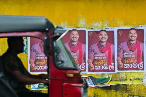 Getty Images A red motorcycle taxi drives past a yellow wall with posters of a man in a red scarf